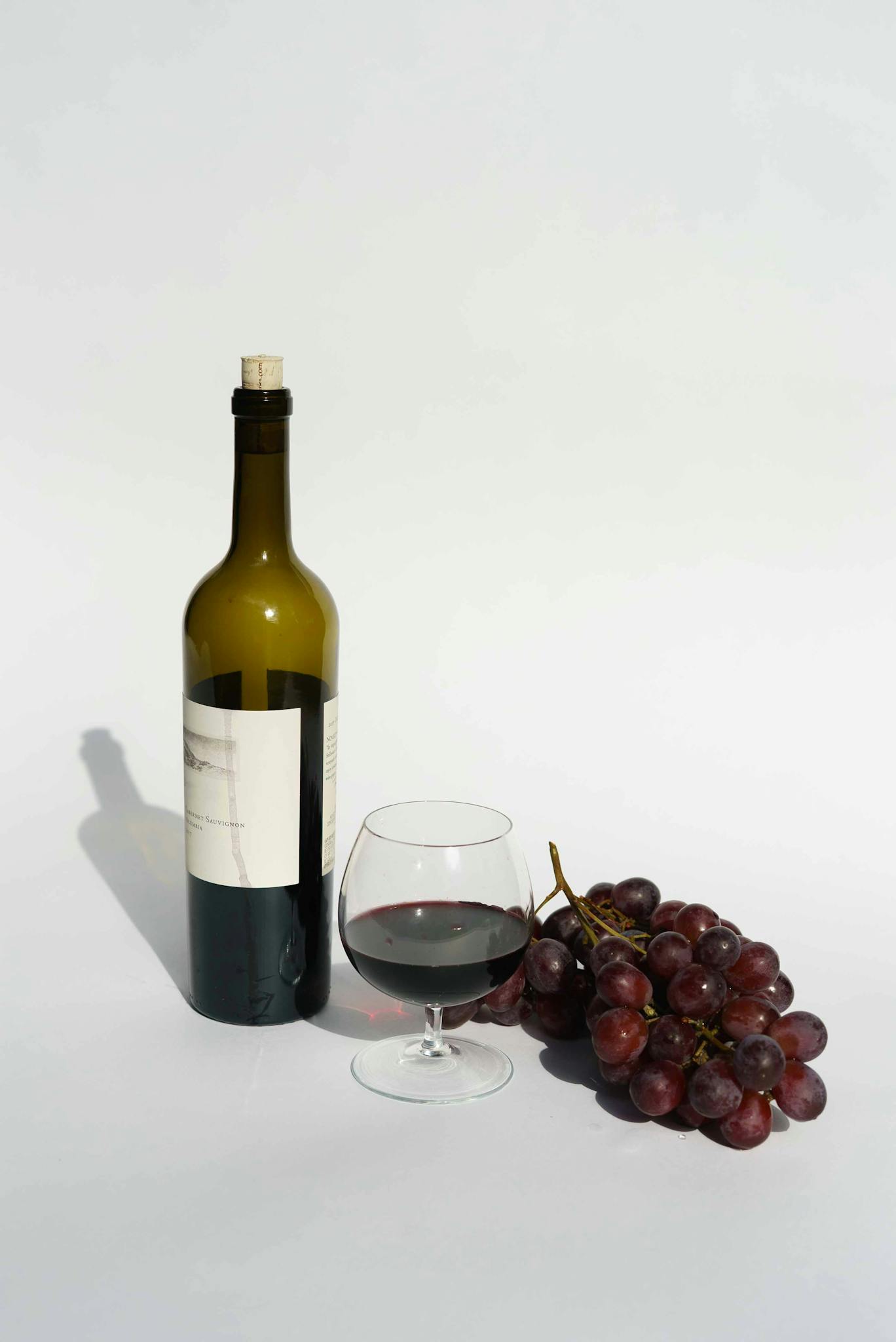 A sophisticated still life featuring red wine, a wine bottle, and fresh grapes on a white background.